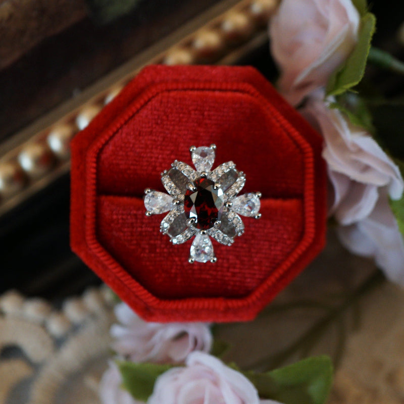 Vintage-style ruby zircon ring featuring an oval deep-red center stone surrounded by pear-cut white zircons arranged like a sparkling gift ribbon, set on an 18K white gold plated band and displayed in a red velvet ring box.