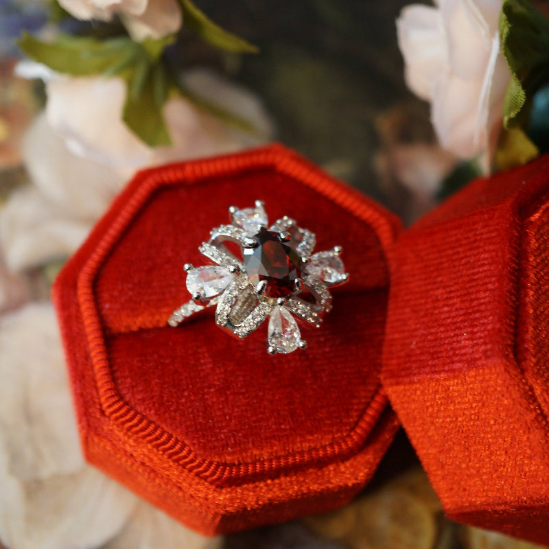 Vintage-style ruby zircon ring featuring an oval deep-red center stone surrounded by pear-cut white zircons arranged like a sparkling gift ribbon, set on an 18K white gold plated band and displayed in a red velvet ring box.