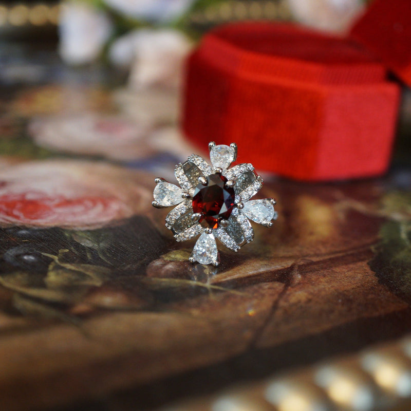 Vintage-style ruby zircon ring featuring an oval deep-red center stone surrounded by pear-cut white zircons arranged like a sparkling gift ribbon, set on an 18K white gold plated band and displayed in a red velvet ring box.