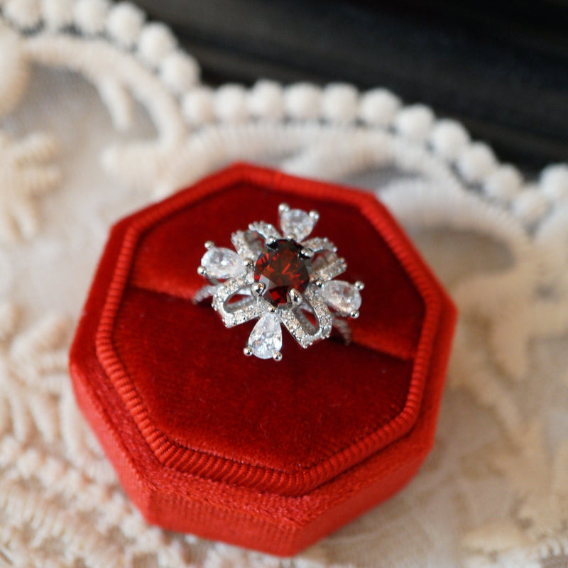 Vintage-style ruby zircon ring featuring an oval deep-red center stone surrounded by pear-cut white zircons arranged like a sparkling gift ribbon, set on an 18K white gold plated band and displayed in a red velvet ring box.
