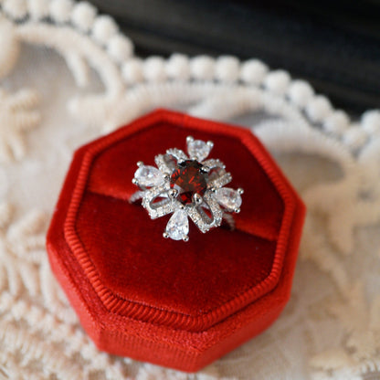 Vintage-style ruby zircon ring featuring an oval deep-red center stone surrounded by pear-cut white zircons arranged like a sparkling gift ribbon, set on an 18K white gold plated band and displayed in a red velvet ring box.