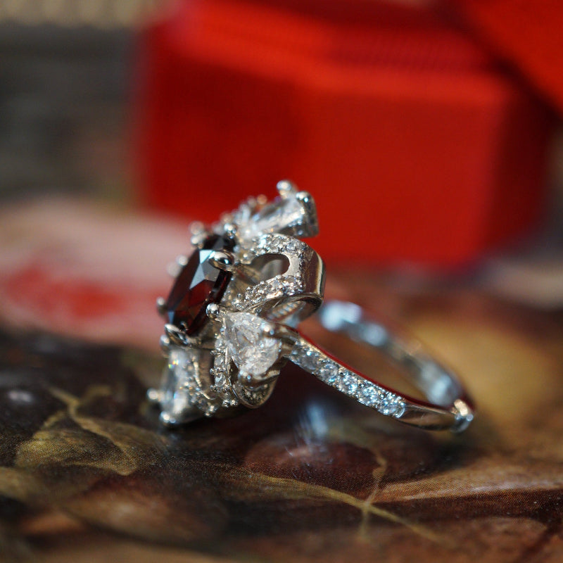 Vintage-style ruby zircon ring featuring an oval deep-red center stone surrounded by pear-cut white zircons arranged like a sparkling gift ribbon, set on an 18K white gold plated band and displayed in a red velvet ring box.