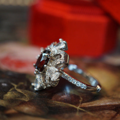 Vintage-style ruby zircon ring featuring an oval deep-red center stone surrounded by pear-cut white zircons arranged like a sparkling gift ribbon, set on an 18K white gold plated band and displayed in a red velvet ring box.
