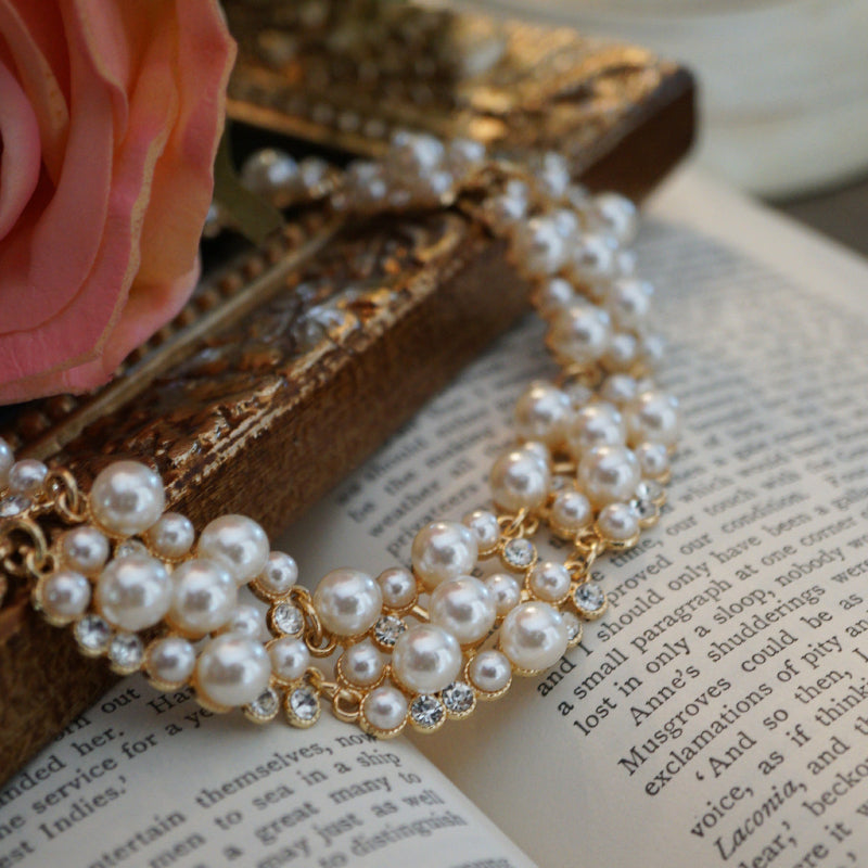A luxurious vintage-style pearl necklace featuring clusters of glossy white pearls and sparkling crystal accents set in gold, displayed elegantly on an ornate gold frame beside soft pink roses and an open book.