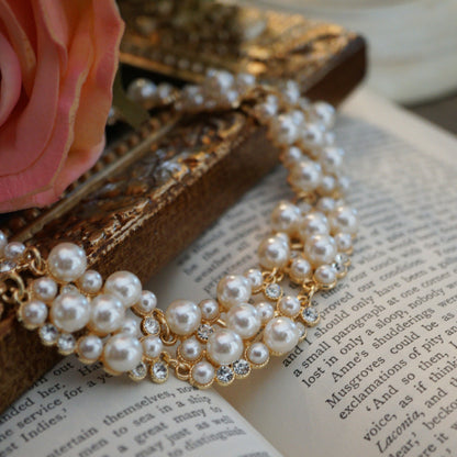 A luxurious vintage-style pearl necklace featuring clusters of glossy white pearls and sparkling crystal accents set in gold, displayed elegantly on an ornate gold frame beside soft pink roses and an open book.