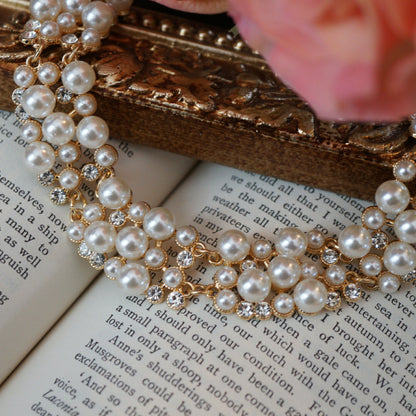 A luxurious vintage-style pearl necklace featuring clusters of glossy white pearls and sparkling crystal accents set in gold, displayed elegantly on an ornate gold frame beside soft pink roses and an open book.