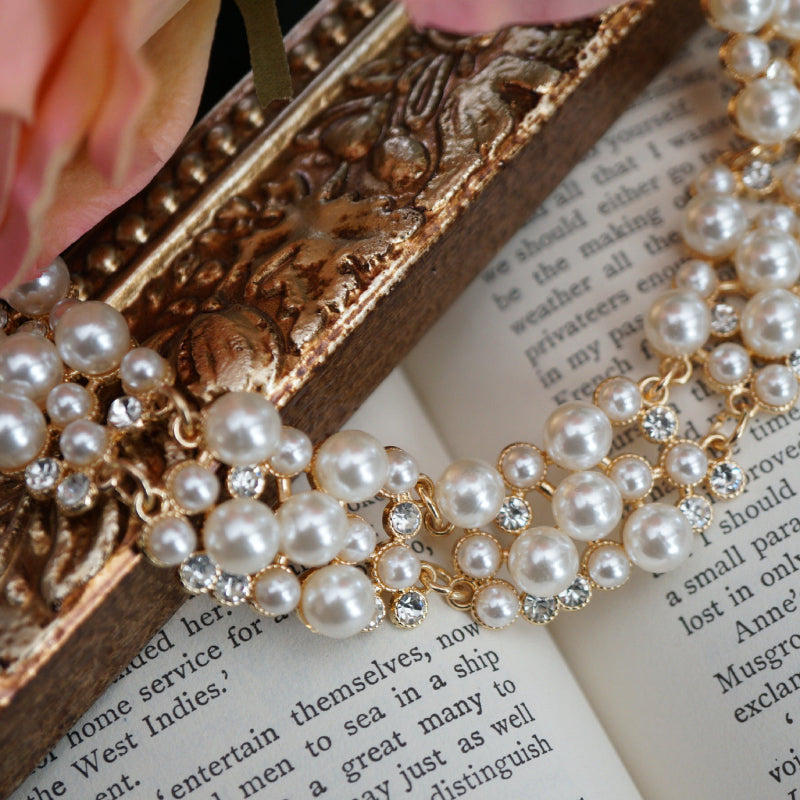 A luxurious vintage-style pearl necklace featuring clusters of glossy white pearls and sparkling crystal accents set in gold, displayed elegantly on an ornate gold frame beside soft pink roses and an open book.