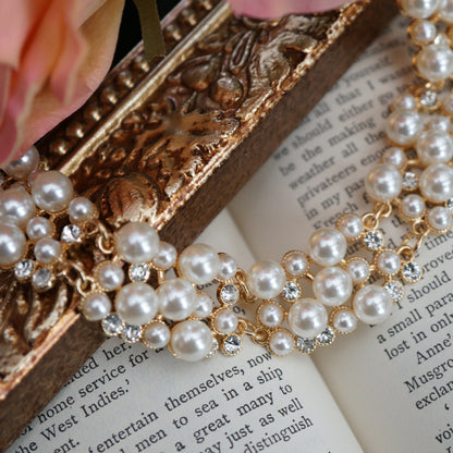 A luxurious vintage-style pearl necklace featuring clusters of glossy white pearls and sparkling crystal accents set in gold, displayed elegantly on an ornate gold frame beside soft pink roses and an open book.