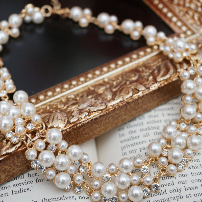 A luxurious vintage-style pearl necklace featuring clusters of glossy white pearls and sparkling crystal accents set in gold, displayed elegantly on an ornate gold frame beside soft pink roses and an open book.