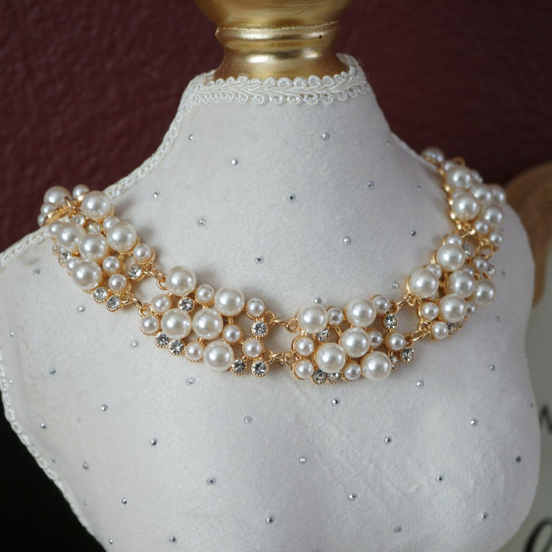 A luxurious vintage-style pearl necklace featuring clusters of glossy white pearls and sparkling crystal accents set in gold, displayed elegantly on an ornate gold frame beside soft pink roses and an open book.